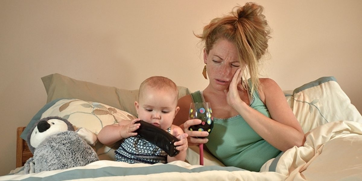 Distraught looking mother laying in bed with her baby, holding a glass of wine.